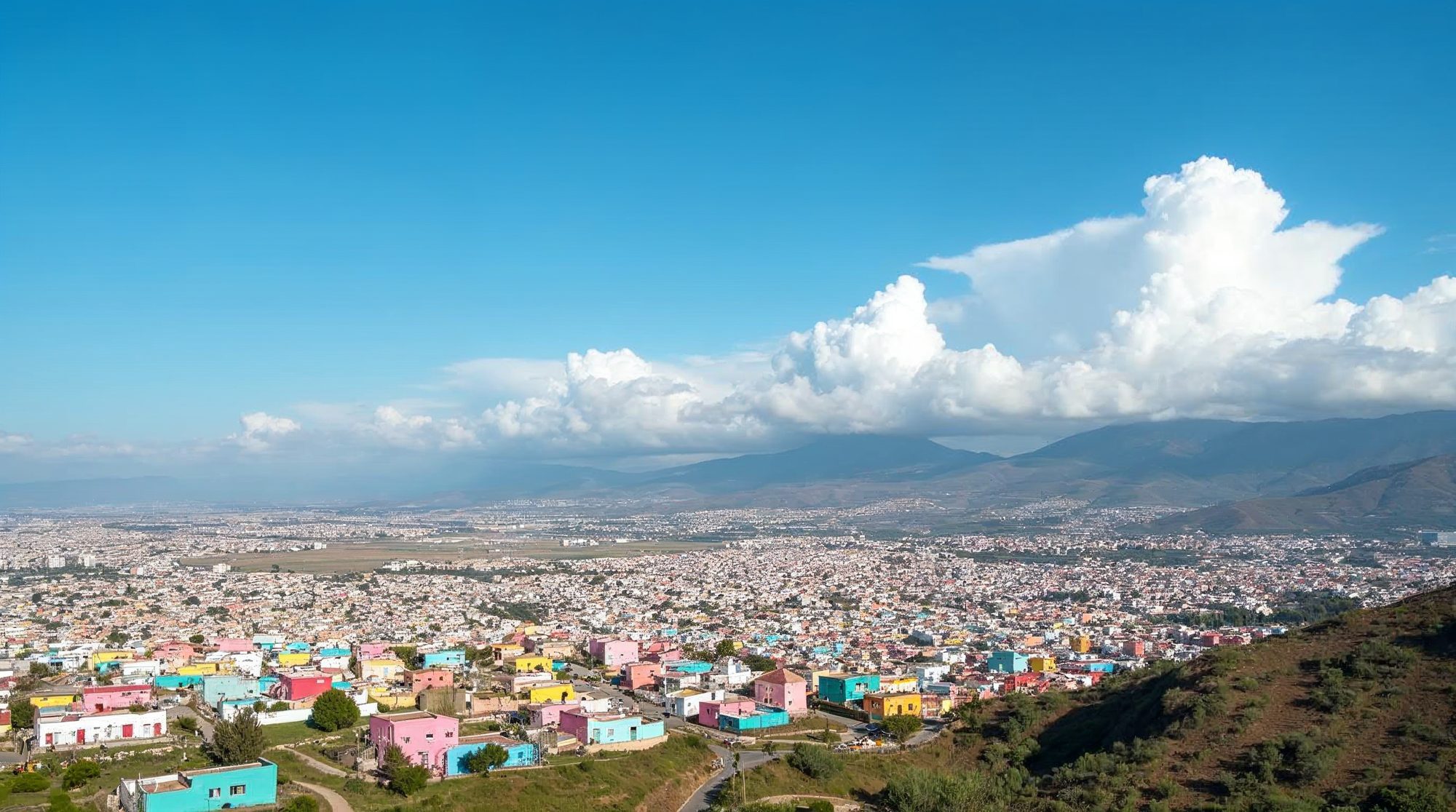 A sunny view over Tijuana, with rows of pink, yellow and turquoise houses spreading across green hills toward distant mountains under a bright blue sky and big white clouds.
